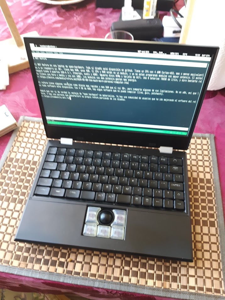 Foto de una reform abierta, la computadora es negra. En la pantalla, un borrador de este articulo. Esta sobre una mesa cuberta con un mantel vino y un "placemat" de bambua cuadriculado.
